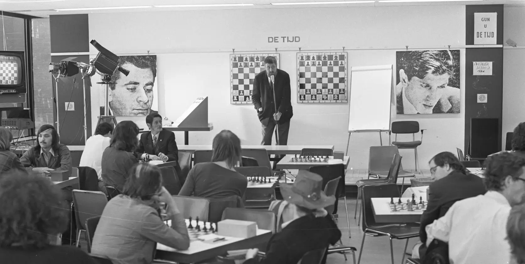 Photo en noir et blanc montrant Jan Hein Donner commentant le championnat du monde d’échecs Fischer-Spassky devant un public, avec les portraits de Boris Spassky et Bobby Fischer affichés derrière lui.
